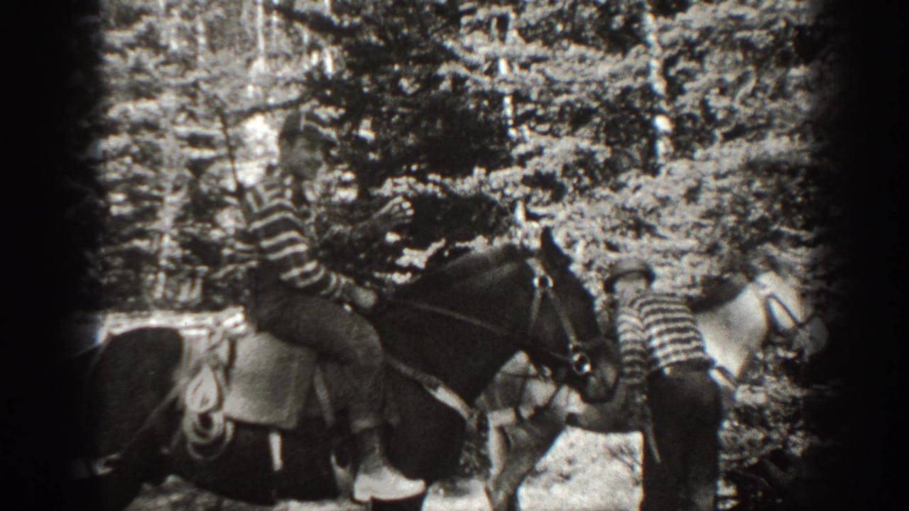 Two riders on saddled horses move through a wooded area.
