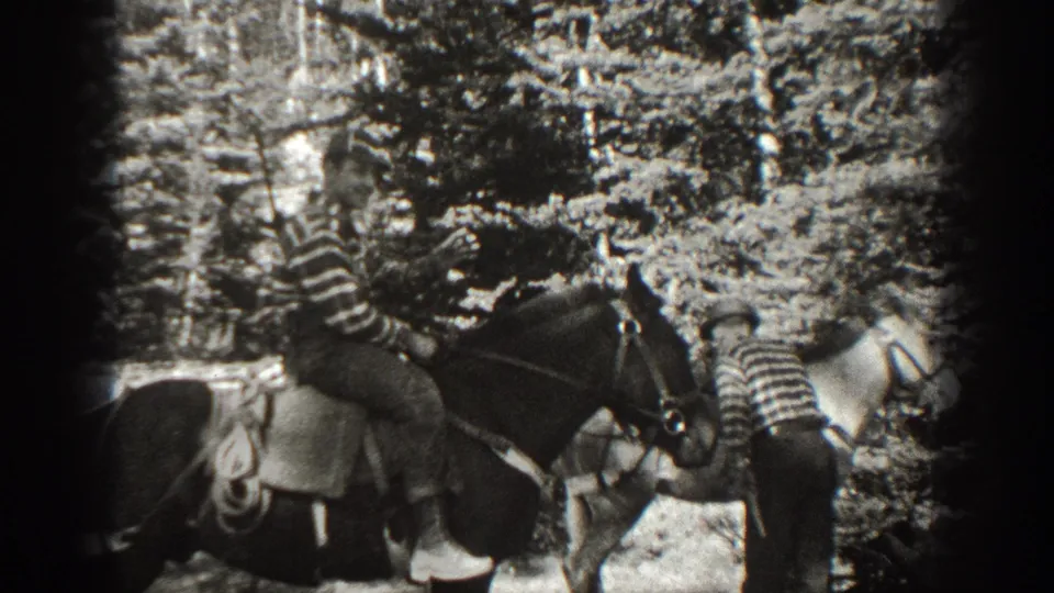 1938 California 8mm frame showing two people in striped shirts riding saddled horses through dense forest growth.