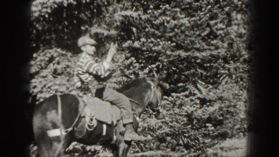 The adult rider raises an open hand from the saddle, with the wooded California setting visible behind him.