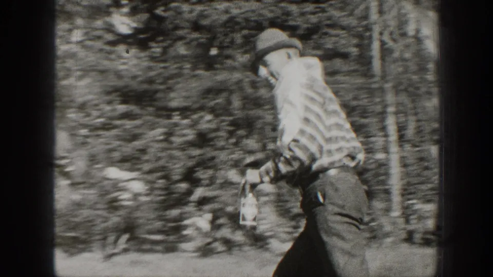 A hat-wearing man in a striped shirt moves across a wooded California setting with a bottle in hand.