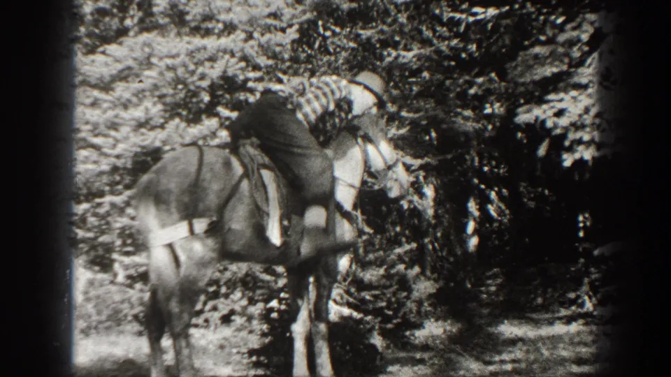 Black-and-white frame of the striped-shirt rider leaning forward on a saddled horse in wooded California footage from 1938.