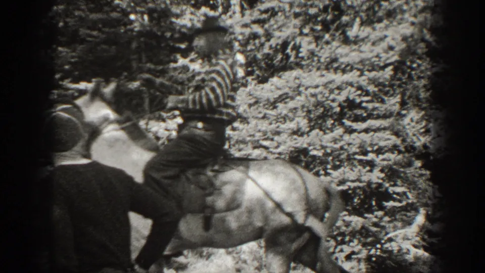 Black-and-white frame showing a second person near the horse as the striped-shirt rider remains mounted in a wooded setting.