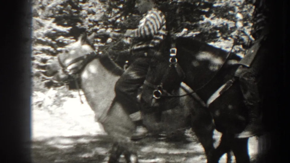 1938 California home-movie frame with men on saddled horses carrying rifles beside trees and rocky terrain.