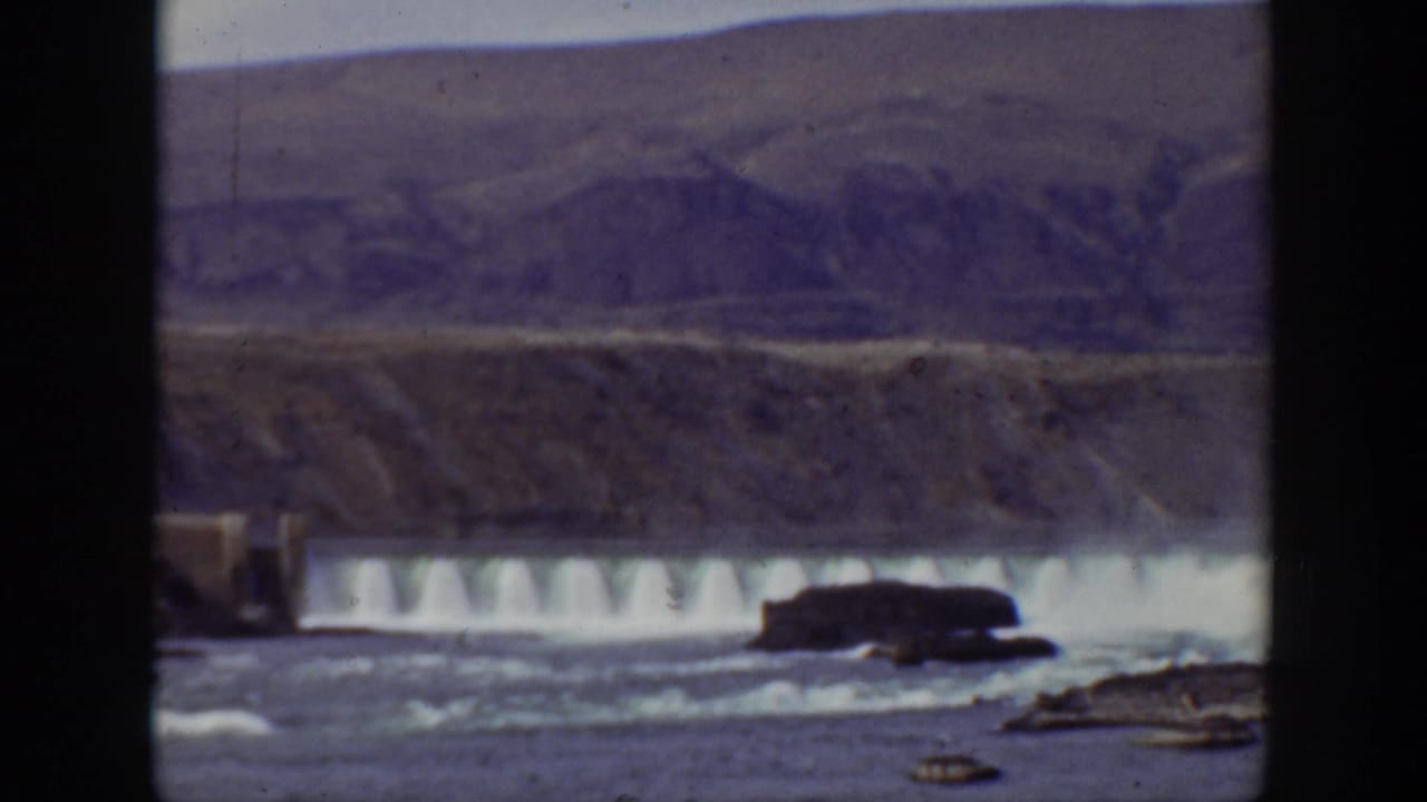 Wide river water cascading over a dam with rocky banks and hills beyond.
