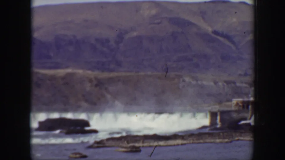 1938 California 8mm frame with soft focus on cascading dam water, rocky foreground, a dark outcrop, and a small structure at the right edge.
