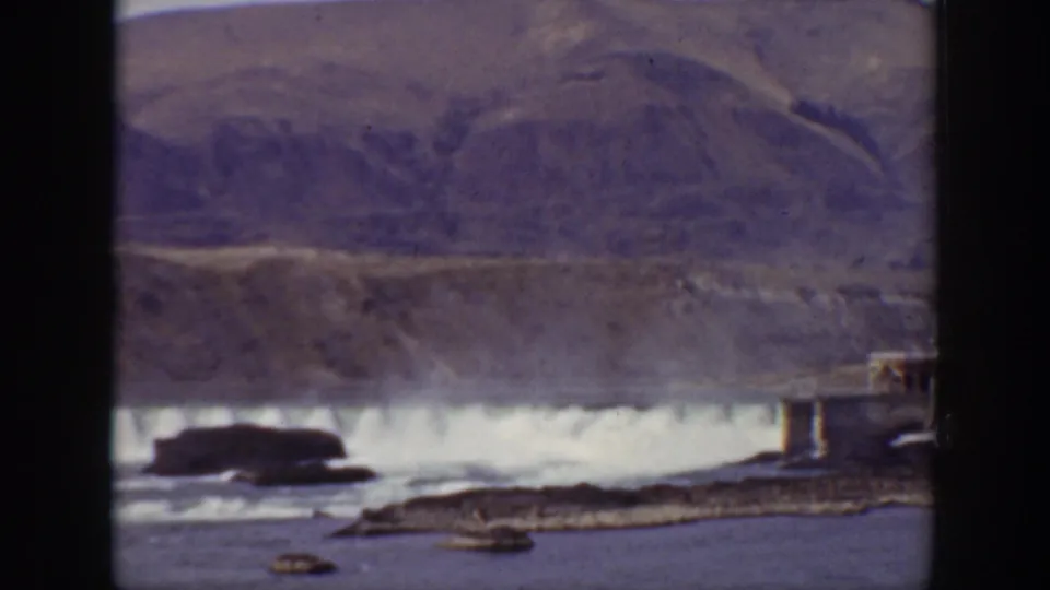 1938 California 8mm frame showing a dam spillway across a river, rocky foreground bars, dark rocks, and muted hills behind.
