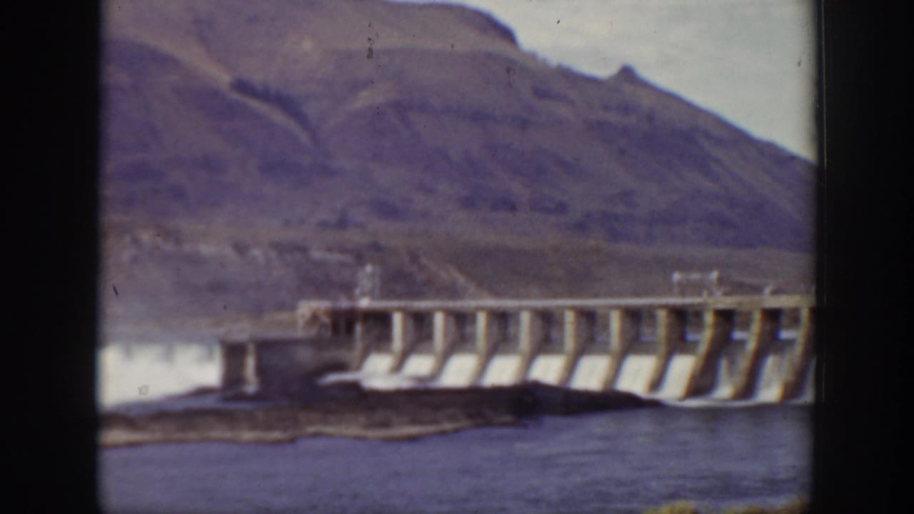 1938 California home-movie frame showing a large concrete dam across water with rugged hills in the background.