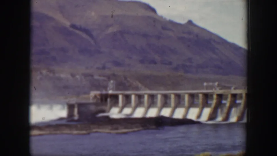 1938 California home-movie frame showing a large concrete dam across water with rugged hills in the background.