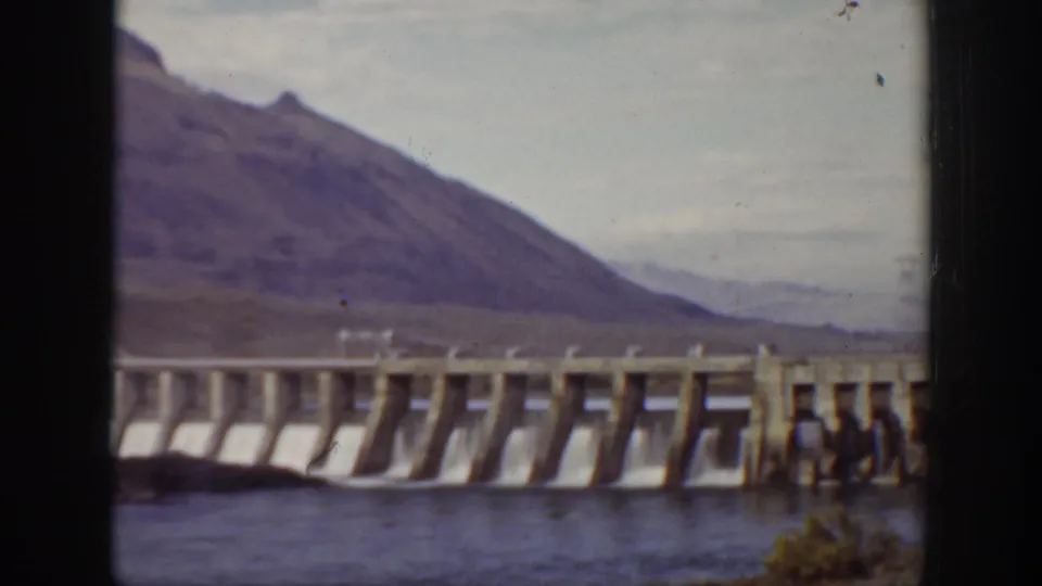Wide 8mm view of the dam from a nearby vantage point, with repeated concrete bays and distant mountain terrain.