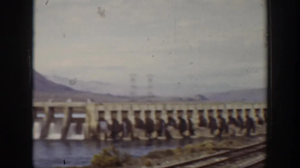 Visitor-shot 1938 California view of the dam with foreground lines, brush, water, and hazy mountains beyond.