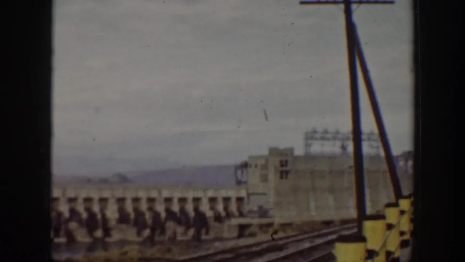 Wide home-movie frame of the dam, an industrial-looking building, utility poles, and distant hills in California.