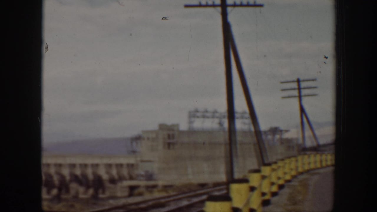1938 California 8mm view from a moving vehicle approaching a large dam, with yellow bridge barriers, utility poles, tracks, and concrete structures.