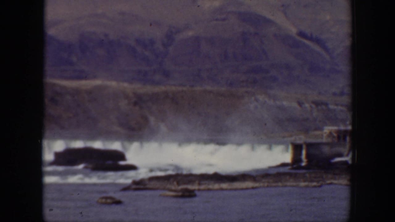Dam spillway with cascading water, rocky river foreground, and mountains behind.