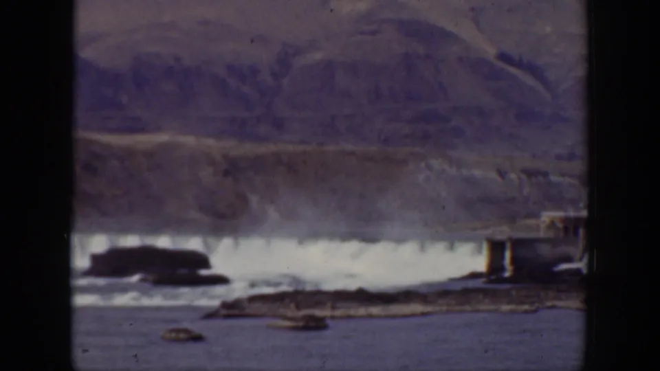 1938 California home-movie frame showing water spilling from a large dam, rocky shoreline in front, industrial edges, and mountains in the distance.