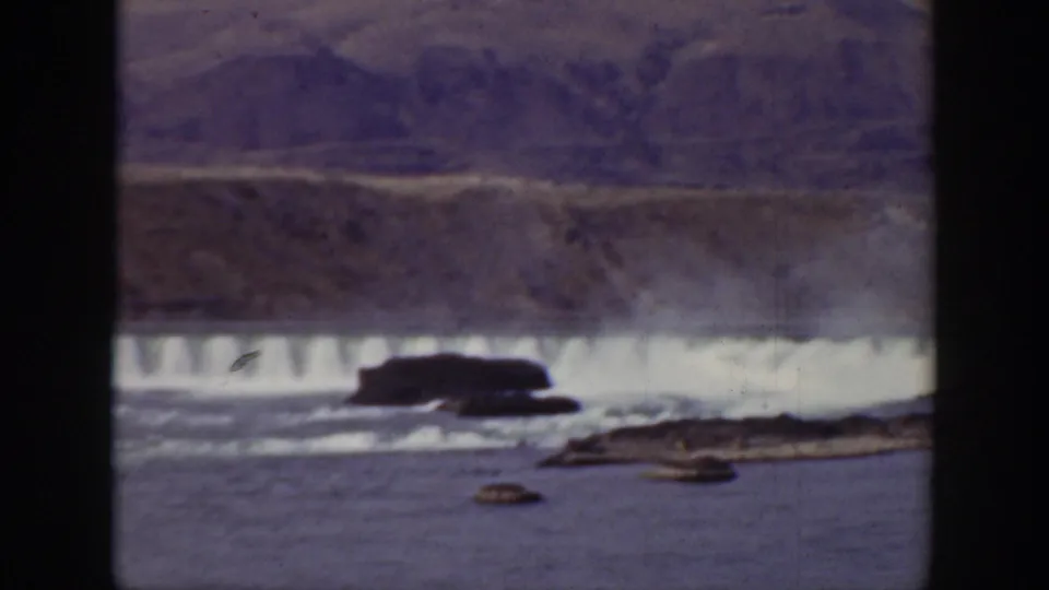 Soft 8mm frame from California in 1938 showing a dam spillway, white water, rocky foreground, and muted mountains beyond.