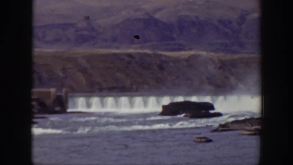 1938 California 8mm view of a dam spillway with white water, rocks in the foreground, a small structure at left, and mountains behind.