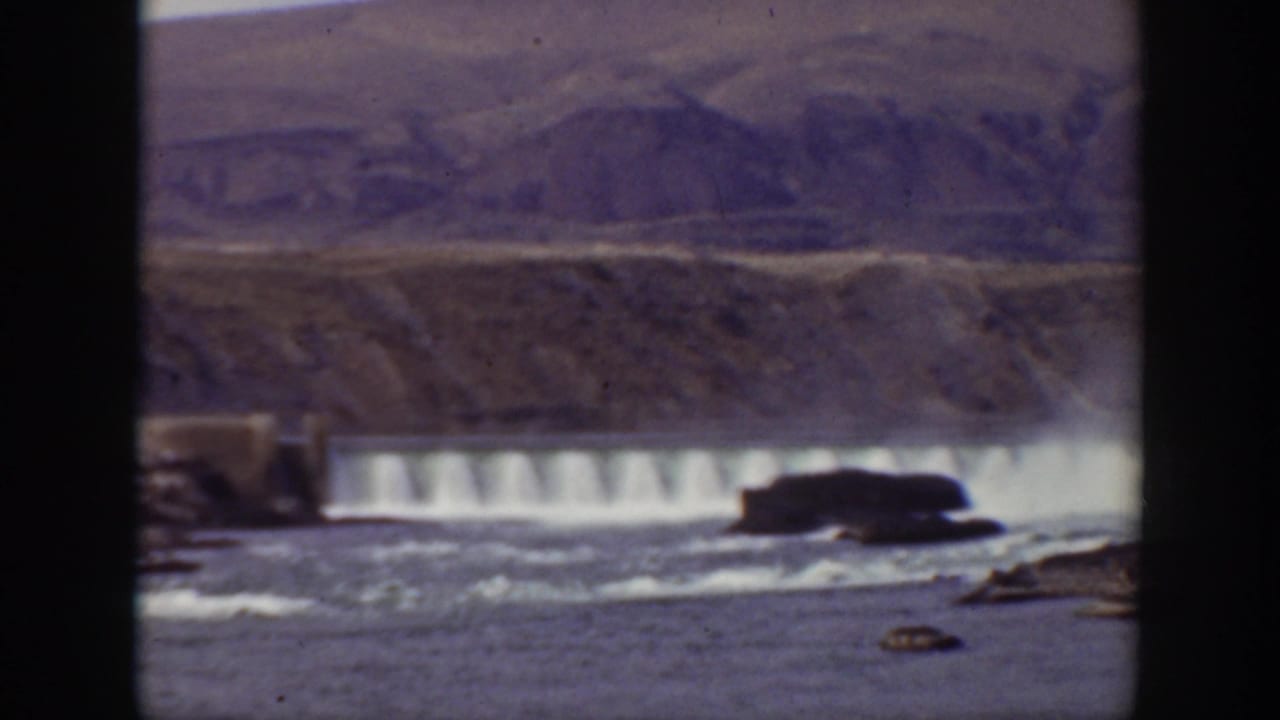 1938 California home-movie frame showing cascading water over a dam with rocky river foreground and dry mountain slopes beyond.