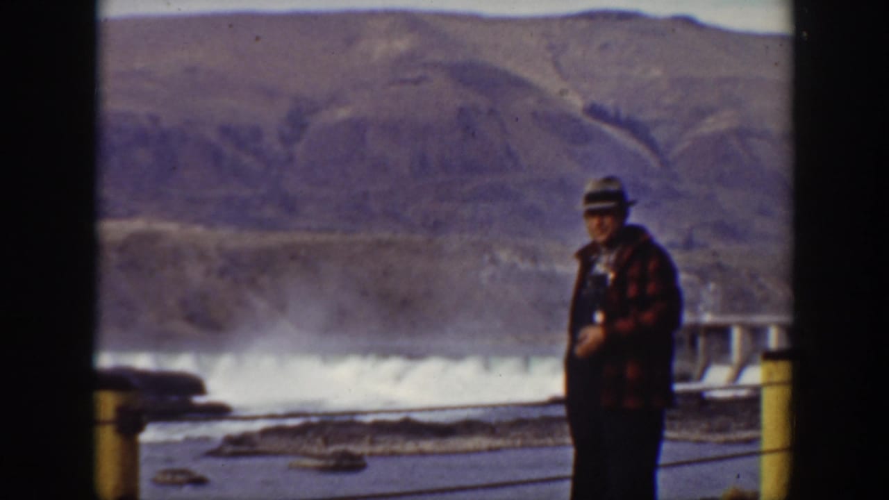 Man in a hat and plaid coat stands near a dam railing.