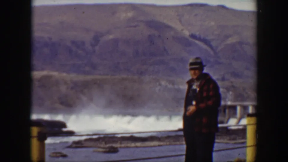 Adult man in a fedora and plaid coat stands near a railing with dam water and California mountain terrain behind him.