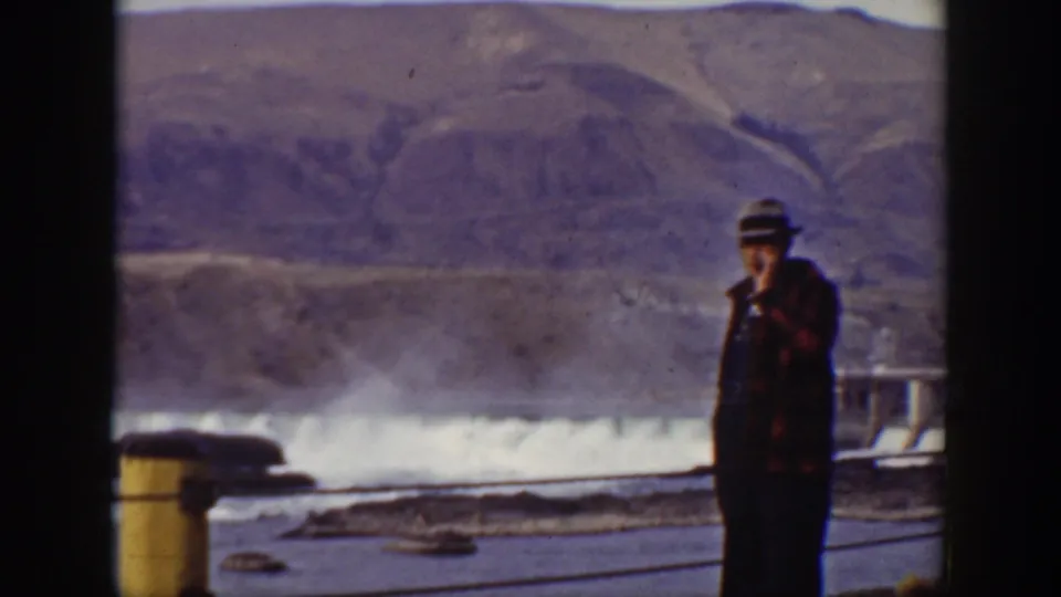 Home-movie frame of the man near the dam, raising a hand toward his face while standing against the spillway and mountain backdrop.