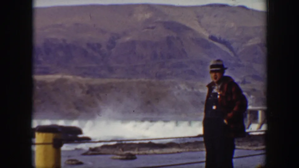 The man in a dark hat and plaid coat stands near the railing with one hand at his side or pocket as dam water flows behind him.