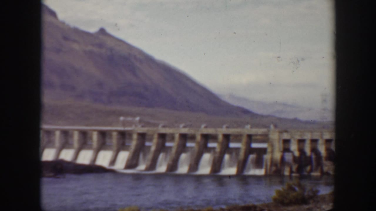 Concrete dam with spillway water below mountains, with a man near the structure.