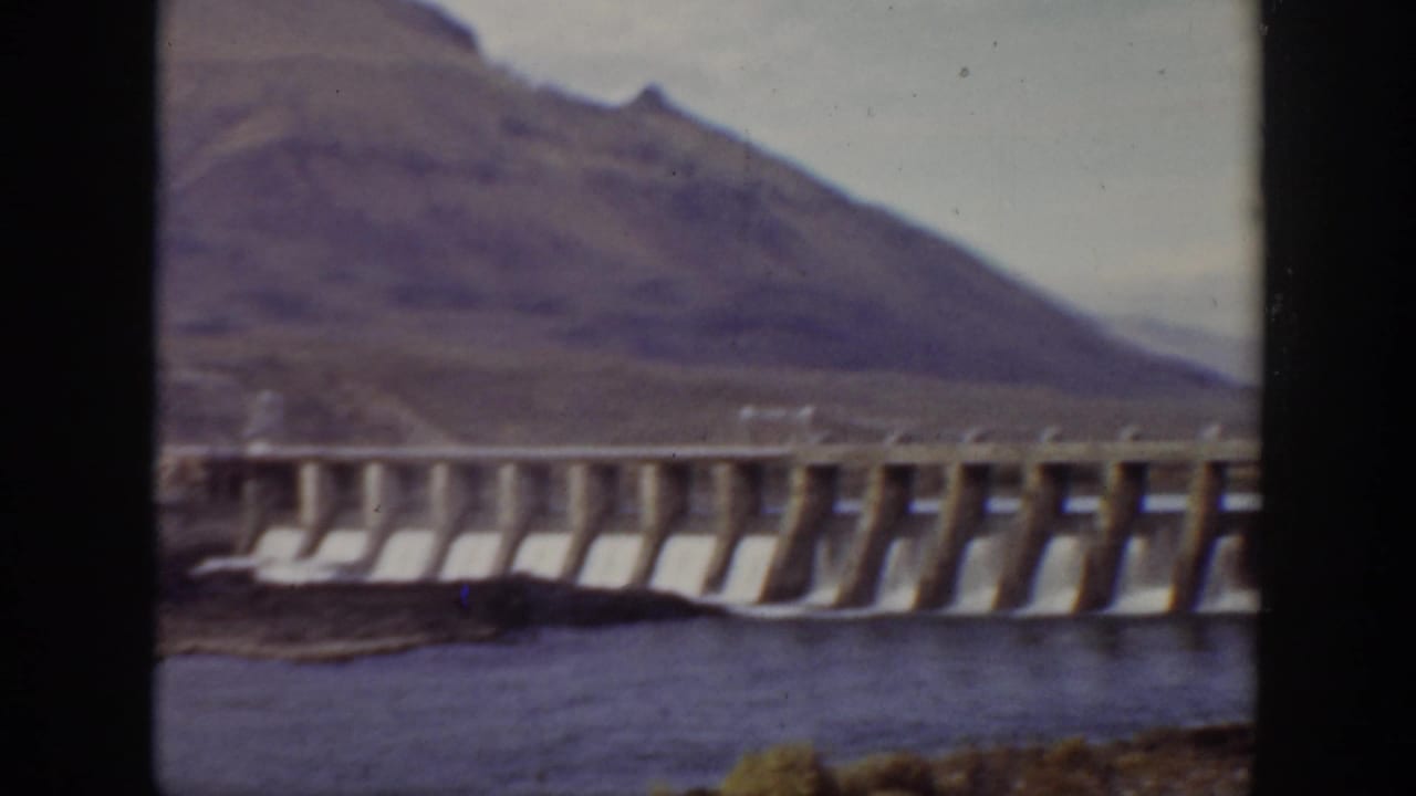 Wide view of a concrete dam across water with mountains in the background.