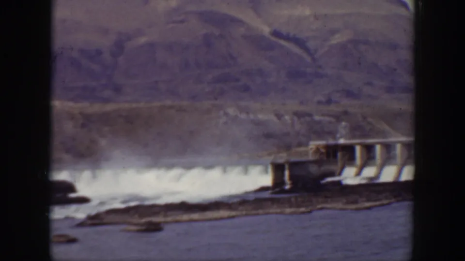 1938 California 8mm frame of white spillway water beside a concrete dam, with river water and mountains beyond.