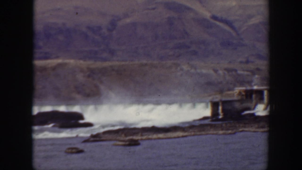 Dam spillway with white water below arid mountains and a rocky river foreground.