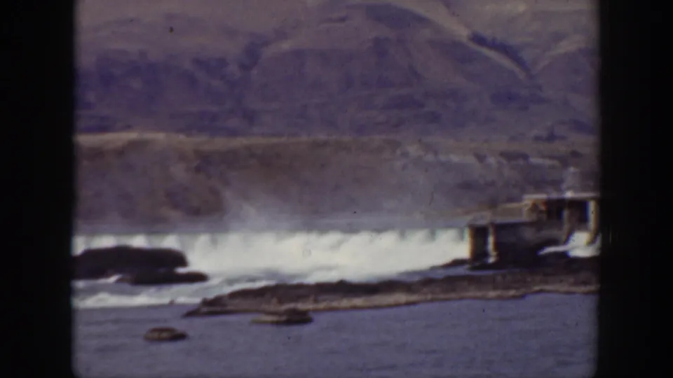 1938 California 8mm frame showing water spilling over a dam in a dry mountain valley with rocks in the river foreground.