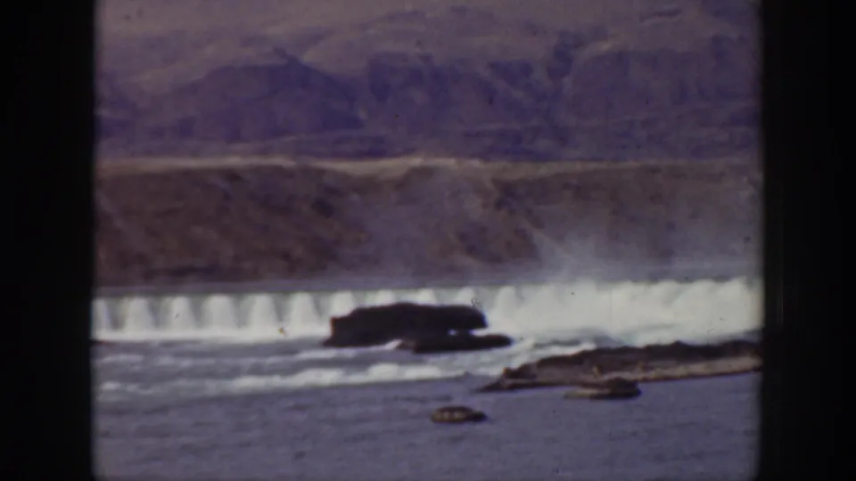1938 California home-movie view of a dam spillway across a river, framed by rocky foreground and dry mountain slopes.