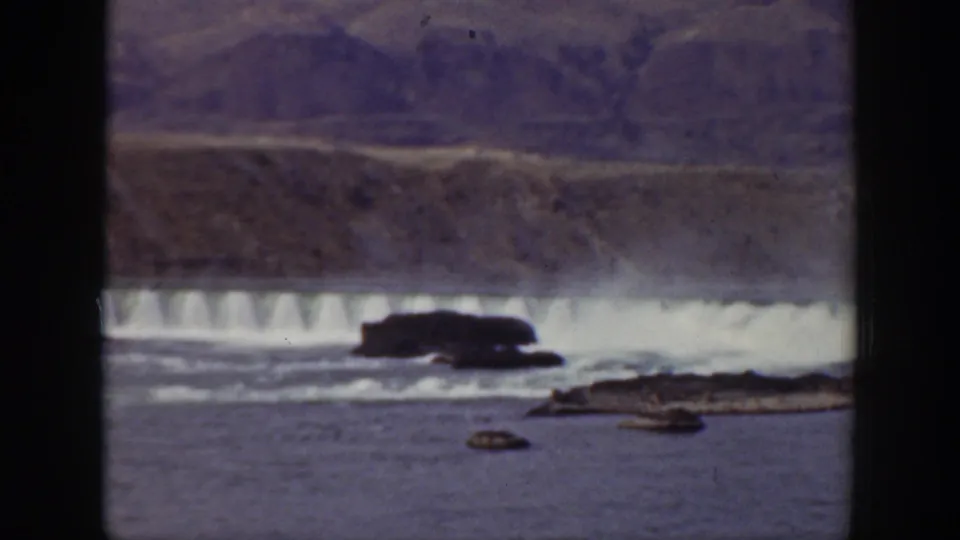1938 California 8mm frame with white water below the dam, dark river rocks, and arid mountains beyond.