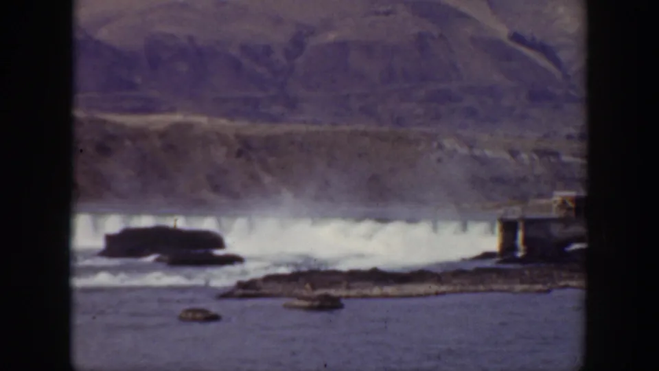 1938 California home-movie frame showing a dam spillway and small structure beside a rocky river in a mountain valley.