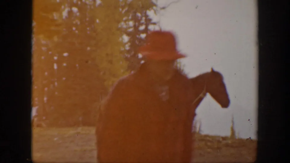 1938 California home-movie frame showing a man in a red hat and plaid jacket leading or walking beside a horse near trees and bright water.