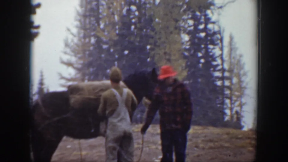 In 1938 California home-movie footage, two men stand near a dark horse in a mountain clearing bordered by tall evergreens.