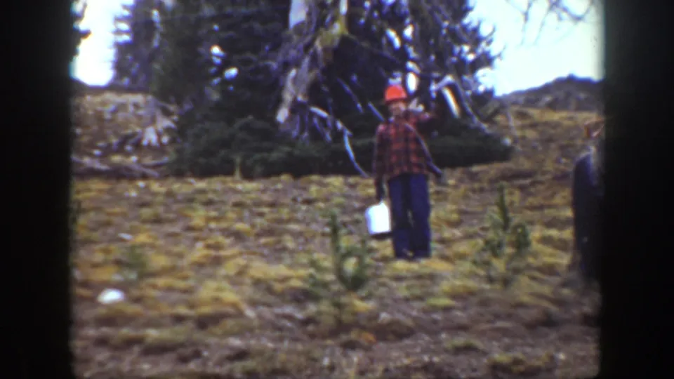 1938 California home-movie frame showing a man in a red hard hat and plaid shirt holding a white bucket near forested hillside terrain.