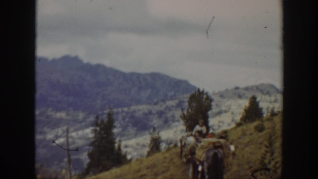 Rider and pack animal farther along the sloping trail with mountains behind.