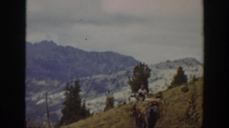 Faded 8mm view of a mounted man and pack animal on a California mountain trail, framed by trees and ridgelines.