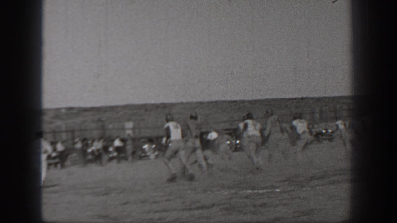 Football players run across an open field with spectators along the far sideline.