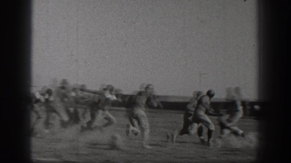Uniformed football players in 1938 Washington footage appear in motion during a running play, with a field barrier and goal posts visible behind them.