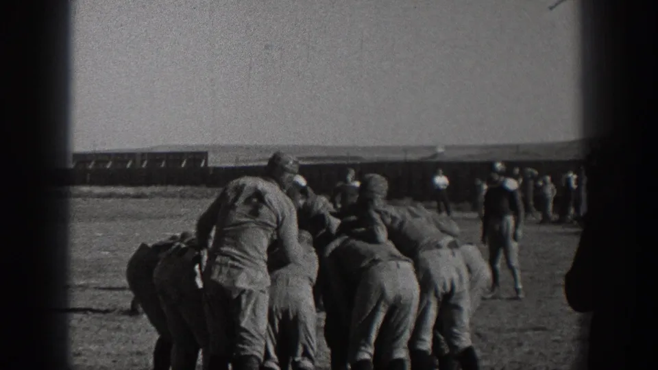 The 1938 Washington home-movie frame shows football players huddled in period uniforms, with a few spectators and the field edge behind them.