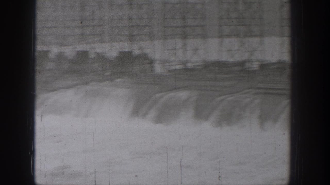 1938 Washington home-movie frame showing turbulent river water beneath a large bridge or spillway, seen through dark vertical divisions.