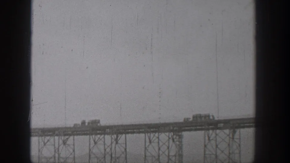 1938 Washington home-movie frame showing trucks or cars moving across a distant elevated steel bridge.