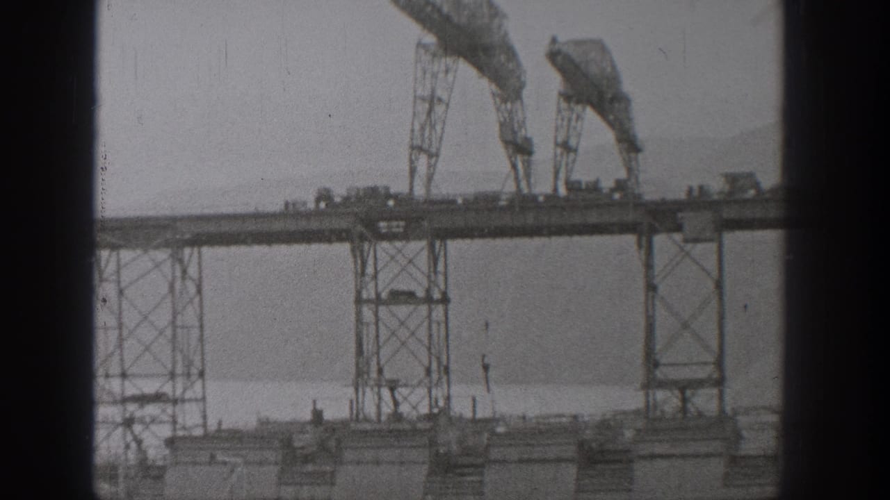 Bridge construction span with cranes, towers, and small vehicles across the deck.