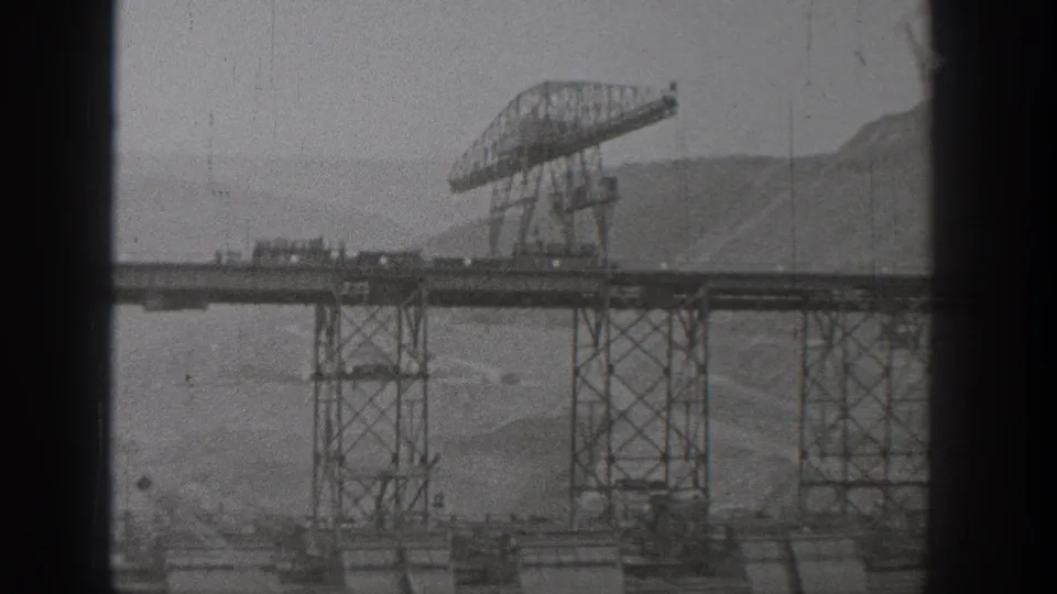 1938 Washington 8mm frame showing a wider bridge construction view with truss work, towers, vehicles, and shoreline terrain.