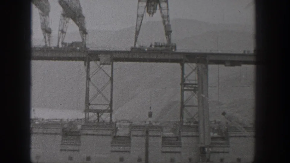 Black-and-white 8mm frame from Washington in 1938 showing bridge construction with cranes, deck sections, supports, and hazy landscape.