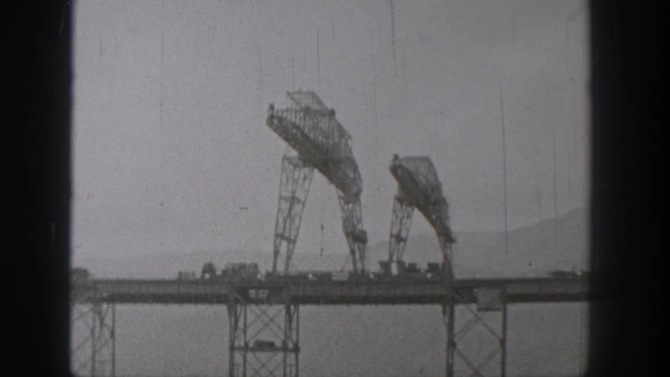 1938 Washington 8mm footage frame showing two large cranes on an incomplete bridge span with hills faintly visible beyond.