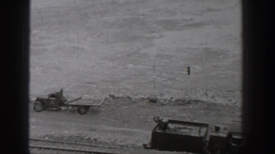 1938 Washington 8mm frame looking down toward a shoreline work area with a small vehicle, flatbed platform, water, and bridge framing.