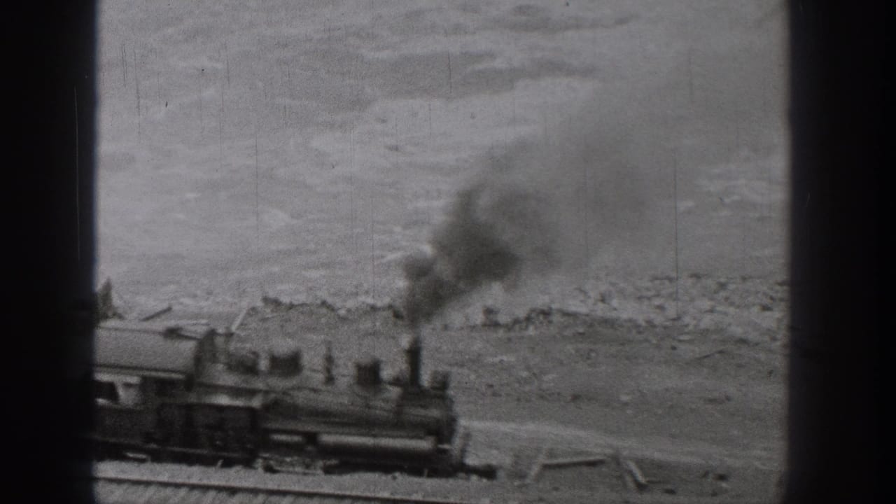 Steam locomotive emits dark smoke while moving beside water and a rocky bank.