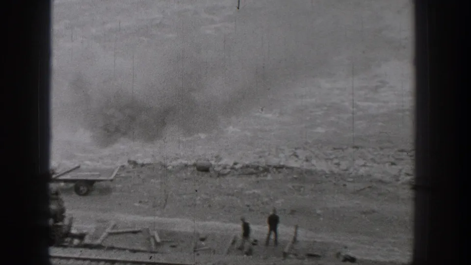 Two distant workers stand near a rail line along the Washington riverbank, with smoke or dust visible over the rough ground.